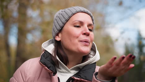 woman in grey beanie and pink jacket blowing dry foliage in her hand, surrounded by blurred autumn trees in the background, creating a peaceful fall atmosphere