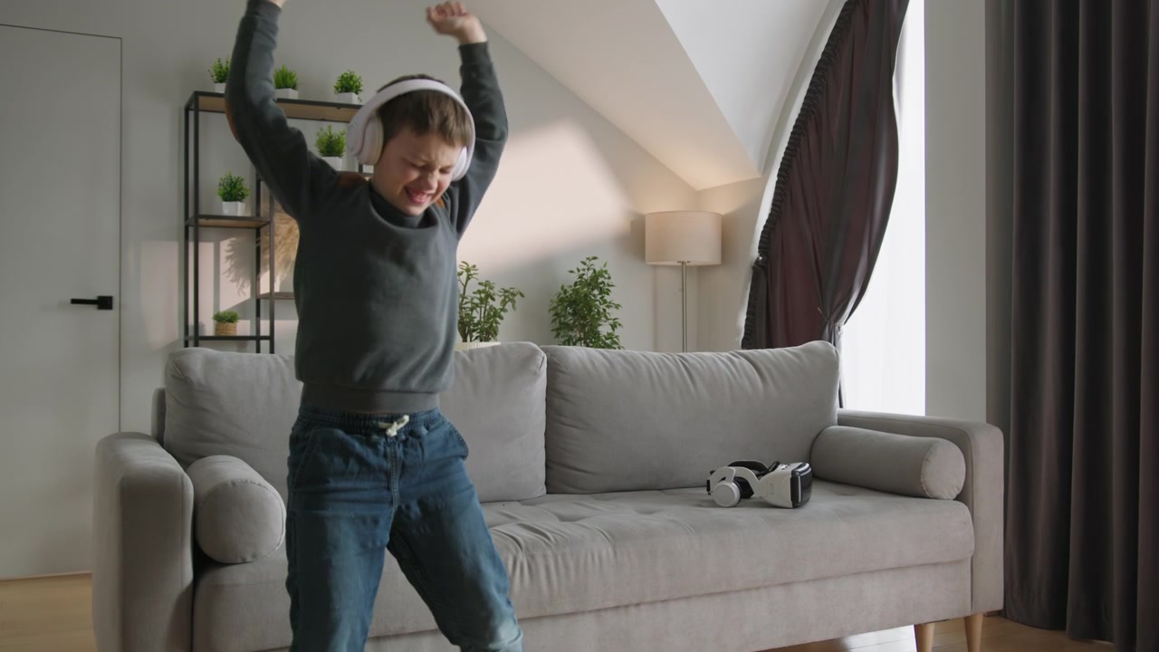Young boy wearing headphones listens to music on his phone while sitting on a comfortable sofa in a