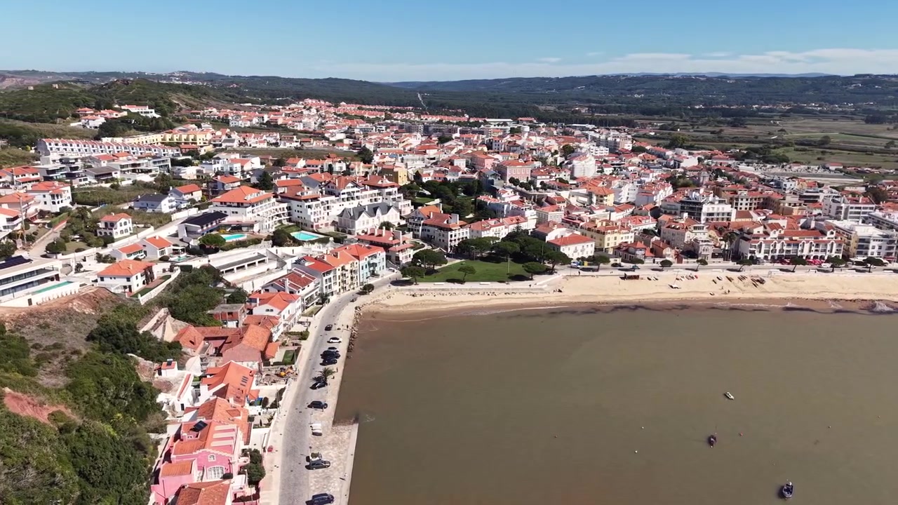 Imagem aérea de drone movendo-se para a direita enquanto orbita sobre a orla marítima de São Martinho do Porto, mostrando a costa arenosa da baía, edifícios à beira-mar e a cidade costeira sob a luz clara da manhã.