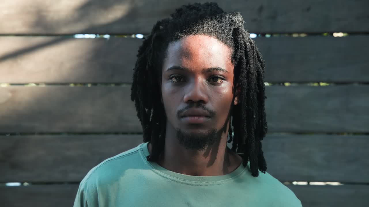 Uomo afroamericano che gira la testa al centro e poi a sinistra davanti a una recinzione di legno, seguendo la luce solare a chiazze