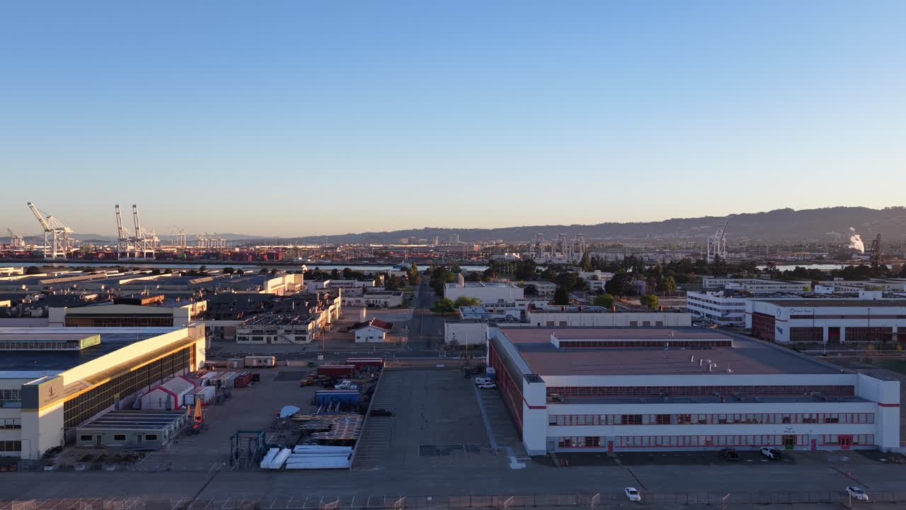 Verfolgende Luftaufnahme über einem Industriepark und Gewerbegebäuden in Alameda, Kalifornien, mit dem Hafen von Oakland im Hintergrund vor einem klaren blauen Himmel bei Sonnenaufgang.