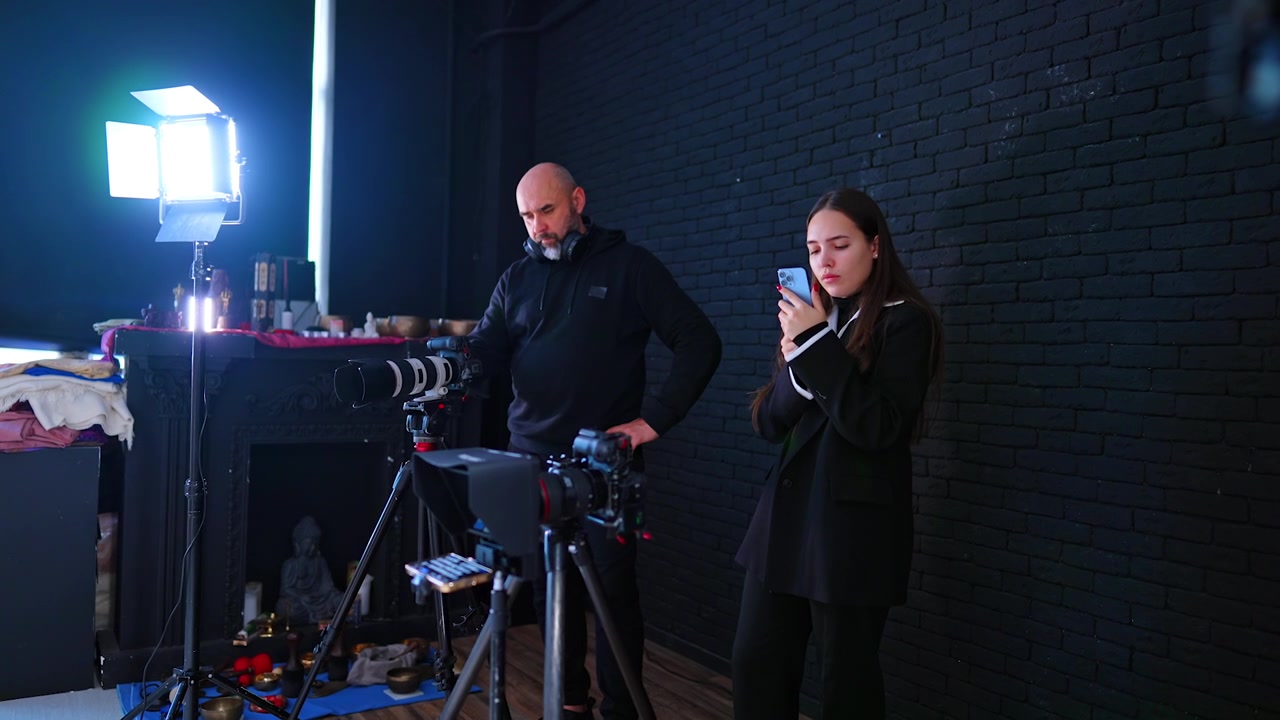 Filmový štáb natáčí rozhovor v tmavém studiu s filmovým osvětlením. Vousatý kameraman a žena prohlížejí telefon vedle kamer na stativech před pozadím z černých cihel.