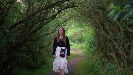 Beautiful-young-woman-with-long-hair-and-a-white-skirt-walks-along-a-romantic-path-in-a-green-tunnel-of-trees-and-bushes