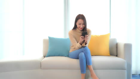 young asian woman sitting on couch and looking at smartphone screen with smile