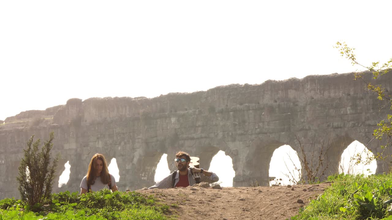 joven pareja encantadora mochileros turistas caminando de la mano hacia el acueducto romano arcos en el parco degli aquedotti ruinas del parque en roma en romántico amanecer brumoso con guitarra y saco de dormir cámara lenta