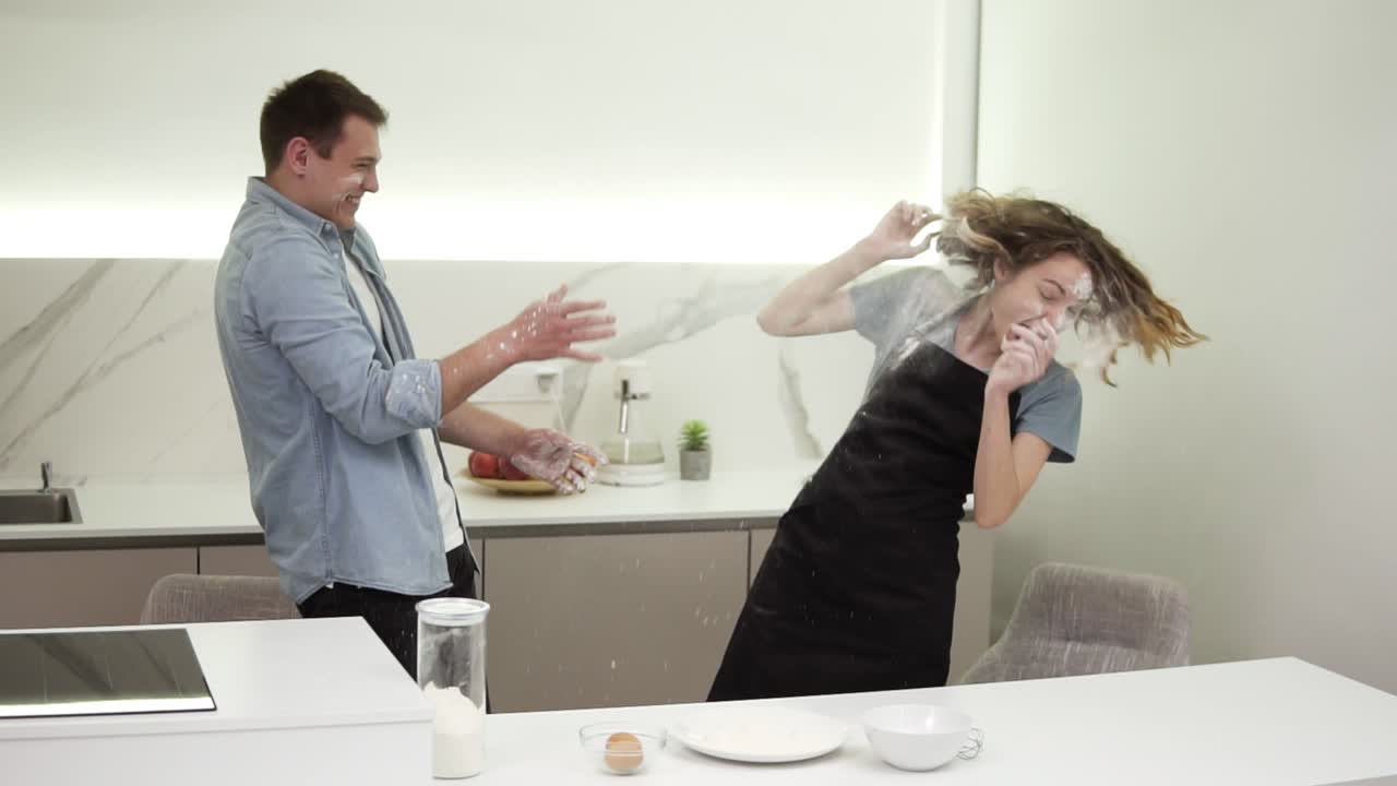 Cute positive girl blowing flour on boyfriend's face in the kitchen, man is throwing back to the face. Happy young couple fooling around while cooking food. Side view, slow motion