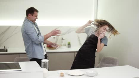 chica linda positiva soplando harina en la cara del novio en la cocina, el hombre está lanzando de nuevo a la cara. feliz pareja joven tonteando mientras cocina comida. vista lateral, cámara lenta