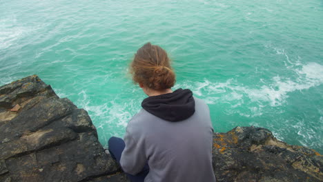 vista trasera de un adolescente varón triste sentado en el borde del acantilado mirando las olas del océano golpeando la roca - ángulo alto