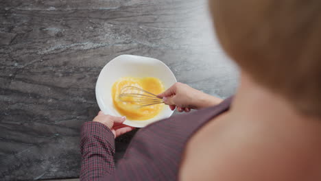 vista trasera de una chica de cabello dorado que lleva un vestido a cuadros moviendo el huevo en un cuenco, ella golpea cuidadosamente la mezcla, creando una textura suave, con un mostrador de cocina limpio y luz natural suave