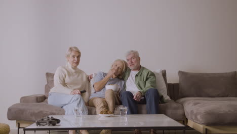 group of three senior people watching tv while sitting on sofa at home