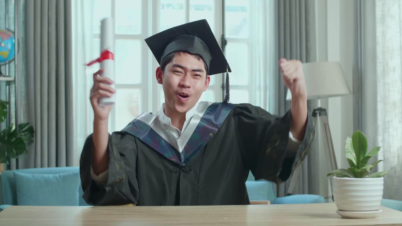 Asian Man Celebrating With University Certificate During An Online Video Call. Male Graduate Wearing A Graduation Gown And Cap Sitting On The Living Room