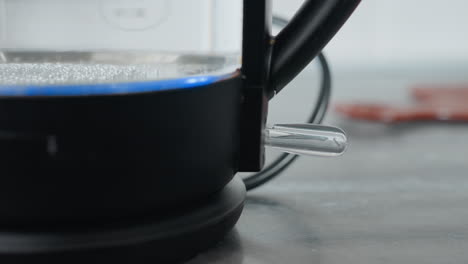 primer plano de la encendido individual de la caldera de agua eléctrica con luz azul parpadeante, mostrando el nivel de agua en el interior, la tetera está encendida y calentando el agua en el entorno de la cocina