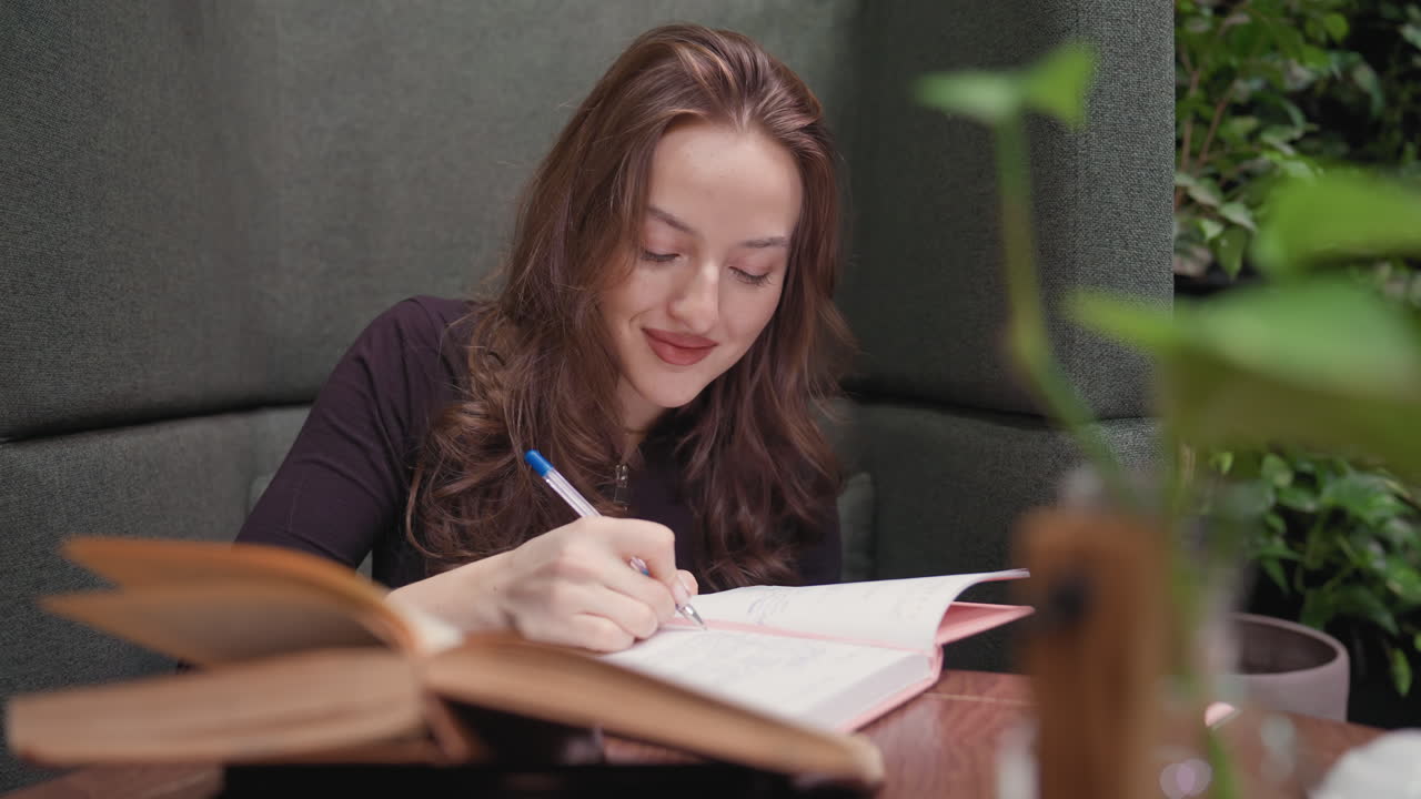 Lady with long brown hair writes in pink notebook while smiling softly, seated in cozy booth with open book nearby and green leafy background adding calm, studious atmosphere to indoor space