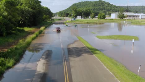 4k-Aufsteigende-Luftaufnahme-Eines-Autos,-Das-Durch-Hochwasser-Fährt-Und-über-Eine-Straße-Außerhalb-Einer-Kleinen-Stadt-Fährt