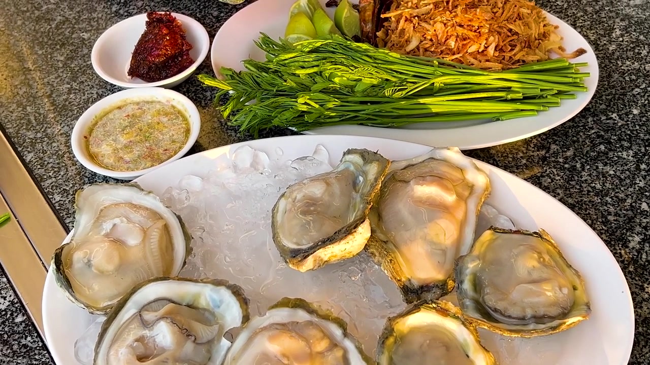 A top-down view of hands preparing fresh raw oysters served with traditional Thai side dishes.
