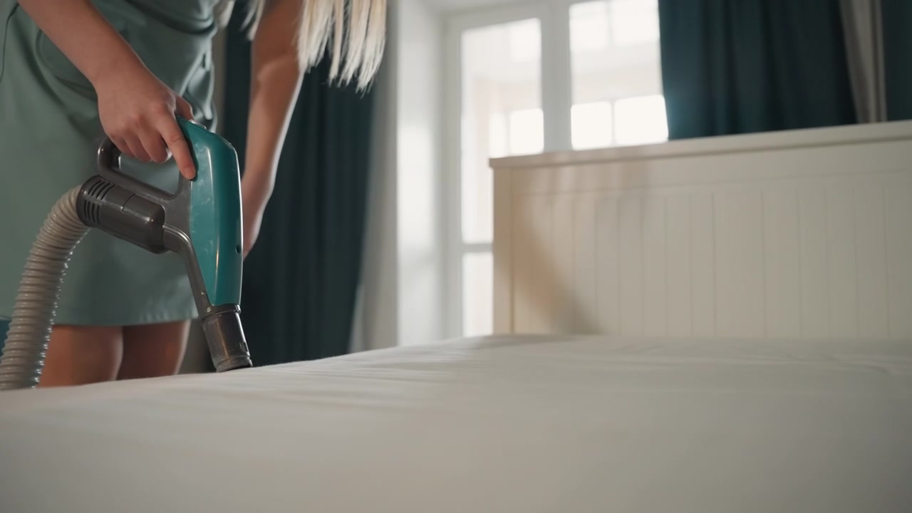 Mulher branca aspirando colchão com ferramenta de estofamento portátil em quarto ensolarado, movimentos lentos e deliberados removendo poeira e cabelo, vestido azul-petróleo e cabelo comprido, cabeceira branca e decoração moderna, focada