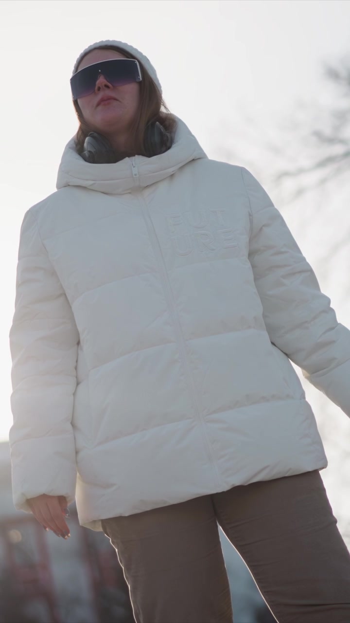 Woman wearing white puffer jacket and beanie holds sunglasses in one hand while adjusting them with other standing outdoors under bright sunlight. background is softly blurred suggesting