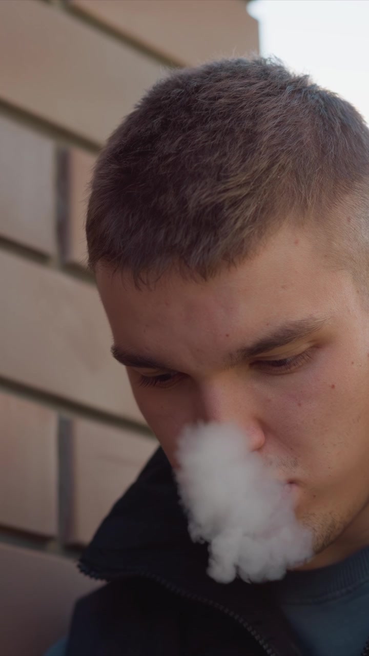 Close up of man holding and using vape device in outdoor setting with soft natural light. subject is focused on vaping interacting closely with object against urban background details
