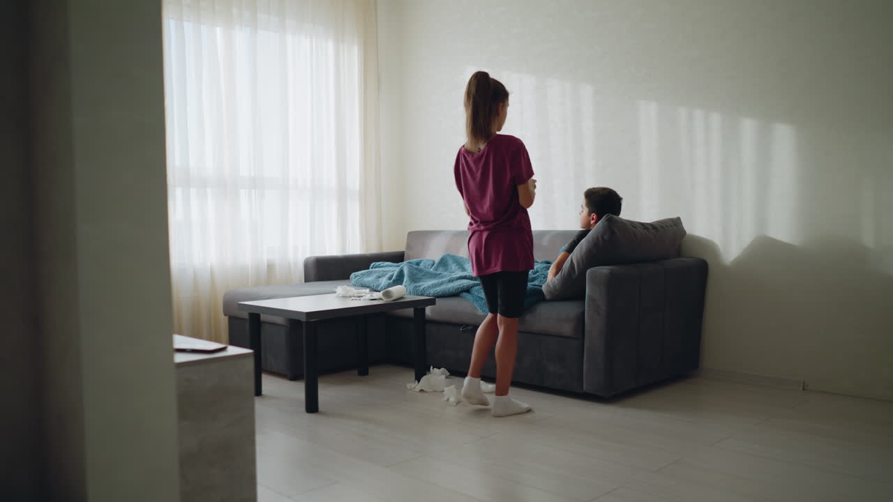 A caring lady serves water to her brother lying sick on the couch while tissue papers are scattered on the floor, highlighting concern and support for a loved one during illness recovery at home