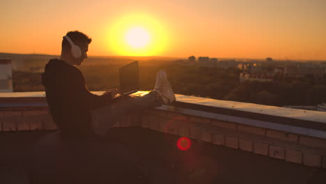 a programmer with a laptop in headphones sits on the roof and writes code at sunset and listens to music. remote work freelancer. freedom to work. typing on a keyboard at sunset with a view of the city.