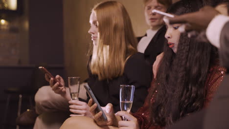 group of friends dressed in stylish clothes sitting on the sofa while using smartphones and drinking champagne during new year eve party