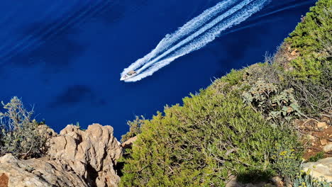 Top-down-drone-flight-above-a-boat-in-calm-waters-of-the-sunny-Navagio-Islands-in-Greece-and-green-plants-with-stones,-slow-motion-and-copy-space