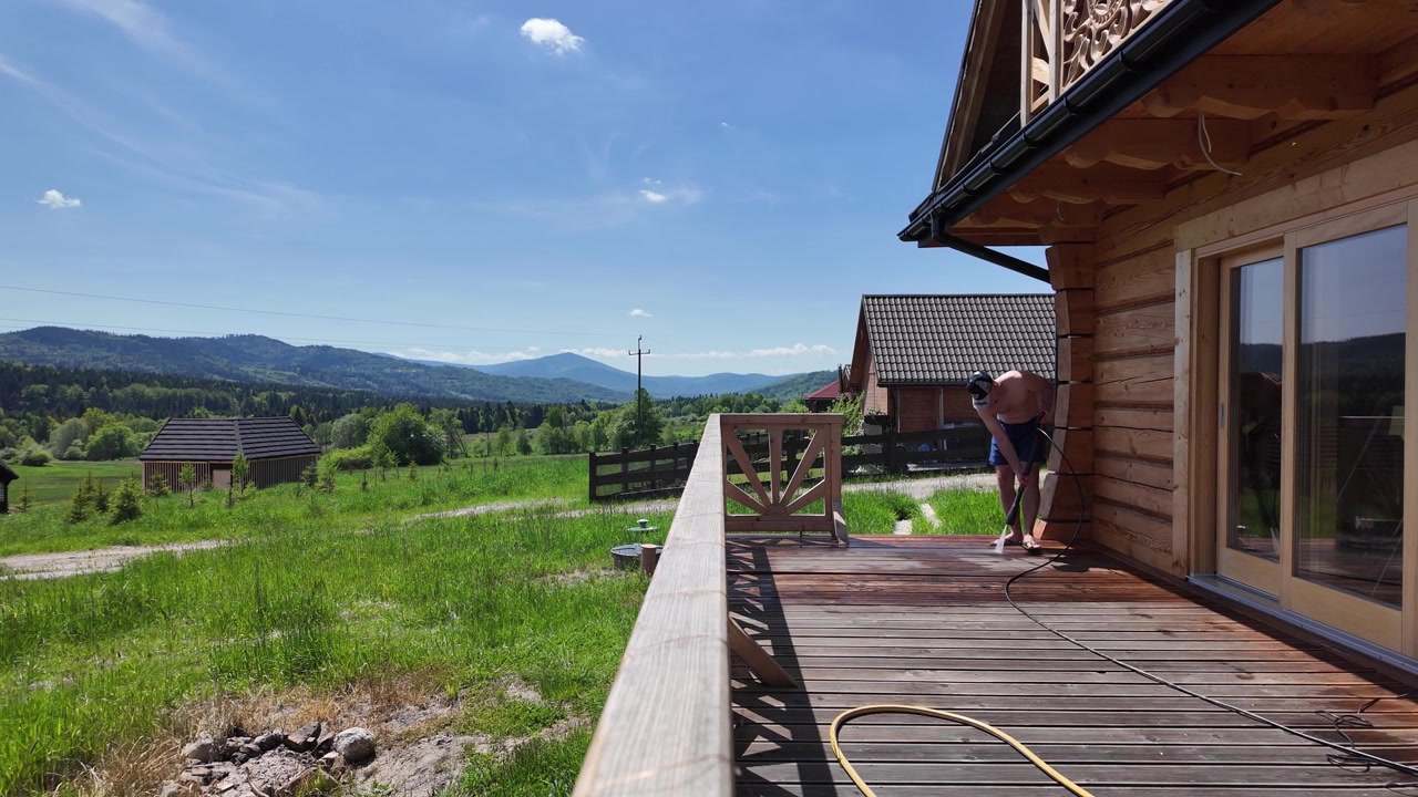Uomo a torso nudo in pantaloncini blu lava a pressione una terrazza in legno presso una baita di tronchi in una soleggiata giornata estiva, circondato da prati verdi, foreste e i monti Beschidi.