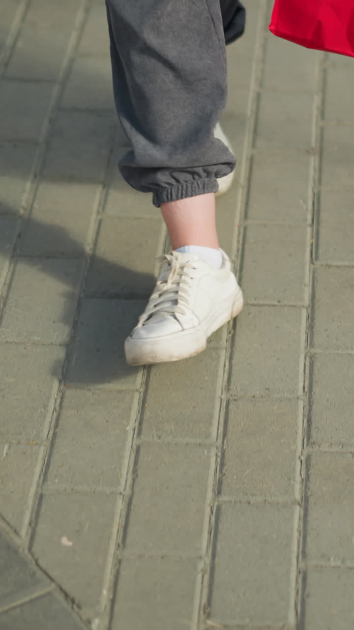 회색 조거와 색 캔버스 신발을 입은 개인의 다리 시각, 녹색, 민트, 빨간색 쇼핑 가방을 들고 간편하게 걸어가는 동안 힌 경로를 걸어 바닥에 작은 종이 조각을 통과