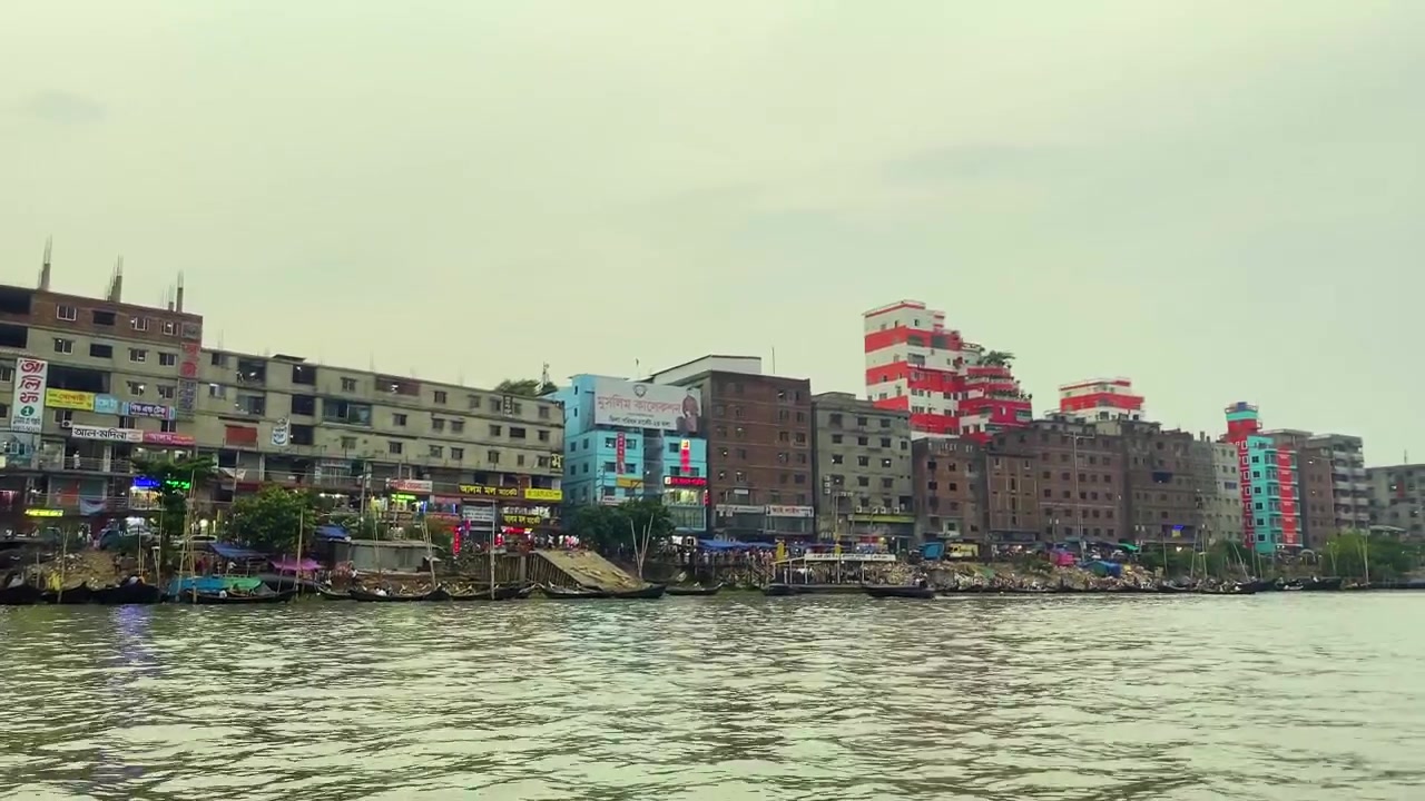 Lungofiume tranquillo del fiume Buriganga con edifici lungo il fiume, molo per barche. Piccola barca a motore che attraversa a Sadarghat, Dhaka, Bangladesh