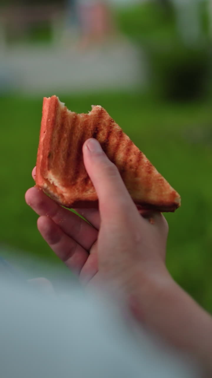 vista trasera de cerca de alguien con una camiseta blanca sosteniendo un sándwich medio comido observándolo con personas y vegetación borrosa en el fondo