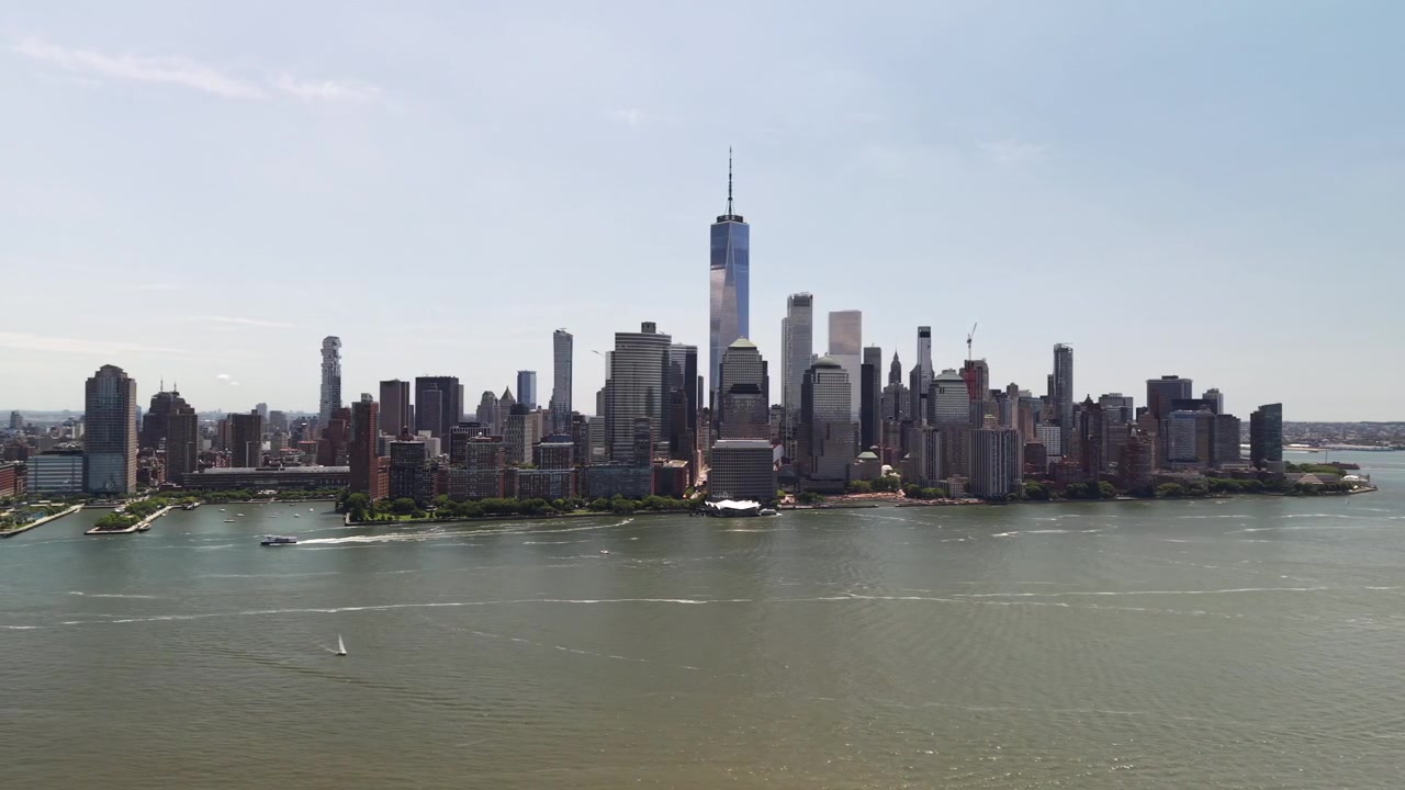 Ripresa aerea con drone che si avvicina lentamente all'iconico skyline di Lower Manhattan, con il One World Trade Center e i grattacieli del distretto finanziario, attraverso il fiume Hudson in una giornata di sole splendente.