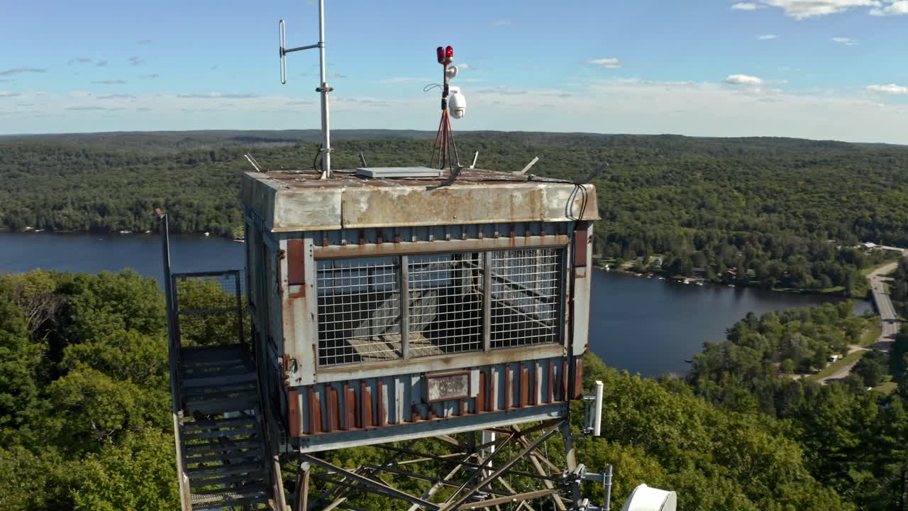 Drone shot rotating around rusty lookout tower will cell equipment overlooking village surrounded by lakes and forested hills