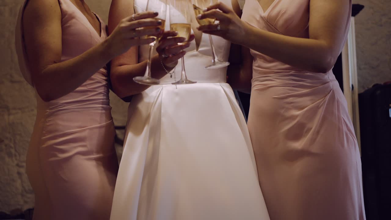 Bride in white gown holding champagne, with bridesmaids in blush dresses embracing