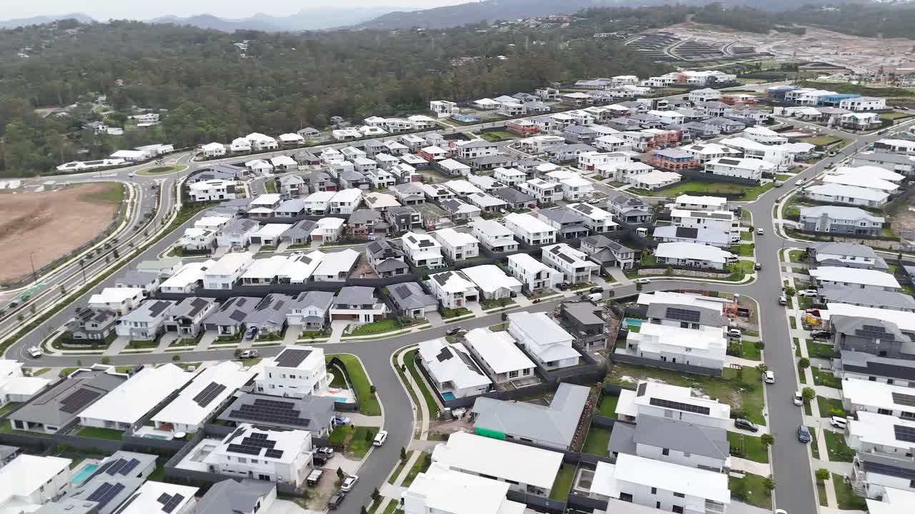 Drone glides above contemporary suburban homes, curving streets, and forested hills under daylight