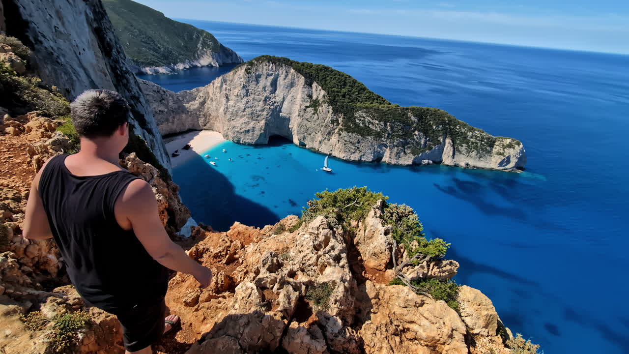 un hombre de pie en un acantilado, mirando hacia abajo en el azul impresionante océano de la isla de navagio, grecia, toma estática con espacio de copia