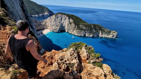 A-man-standing-on-a-cliff,-looking-down-on-the-blue-stunning-ocean-of-the-Navagio-Island,-Greece,-static-shot-with-copy-space