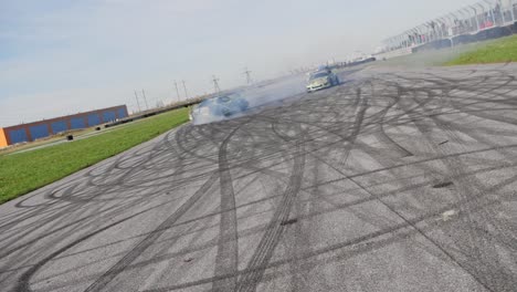 two green muscle drifting cars on a race track doing a parallel drift around each other with deep tire marks on asphalt, slow motion