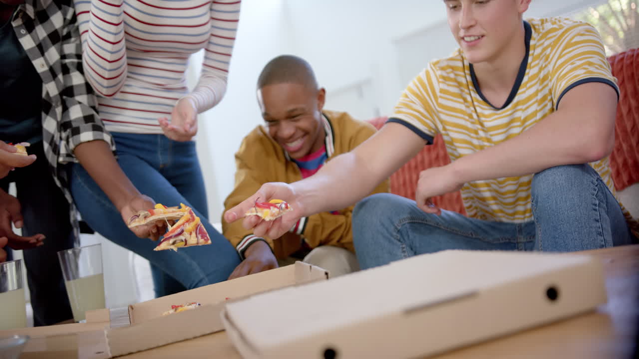 un grupo feliz y diverso de amigos adolescentes sentados en el sofá y comiendo pizza en casa, en cámara lenta