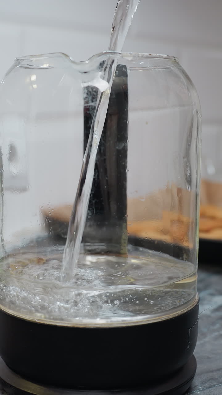 primer plano de agua que se vierte en un frasco de vidrio con un filtro, el fondo borroso presenta un enchufe de pared de la cocina, una bandeja con galletas y una taza