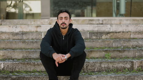 Front-view-of-serious-young-Arabic-handsome-man-with-dark-curly-hair-and-beard-in-black-hoodie-sitting-on-stairs-outside