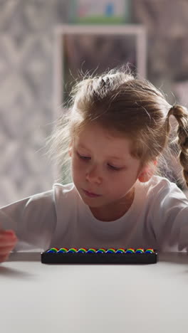 active little girl with colorful toy abacus counts by finger and talks sitting at desk at arithmetic lesson against bookcase closeup slow motion