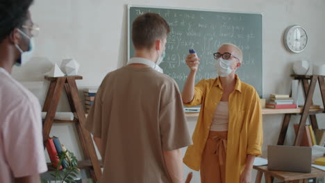 teacher in mask measuring temperature of students in classroom