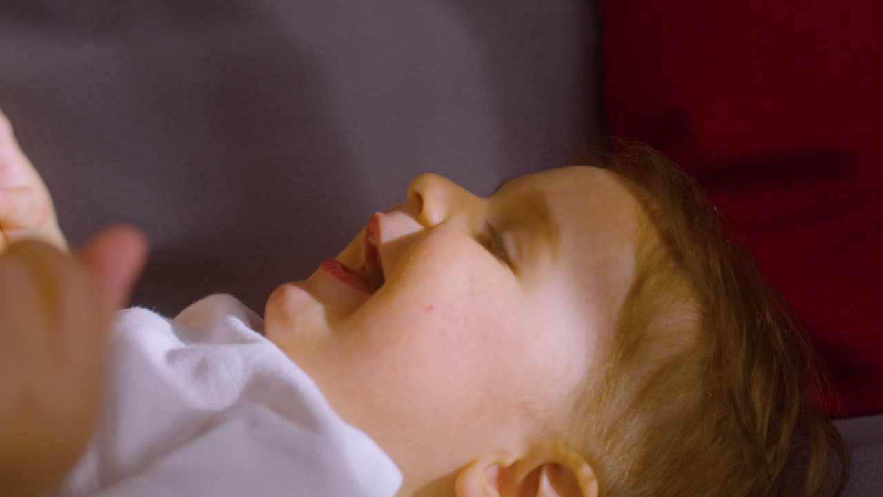 어머니가 간지럽히고 그와 놀고 있는 동안 집에 있는 소파에 누워 웃고 있는 아기의 모습을 가까이서 볼 수 있습니다 2