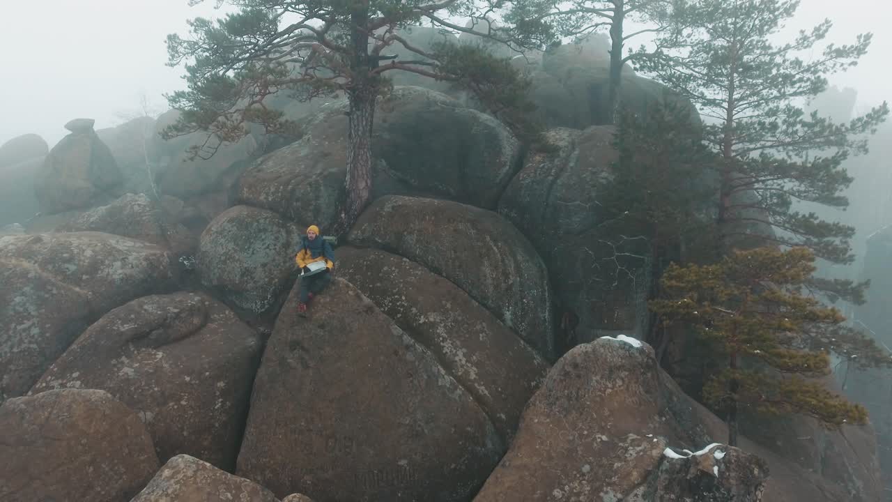 Man working on a laptop on top of rock formations in a foggy landscape