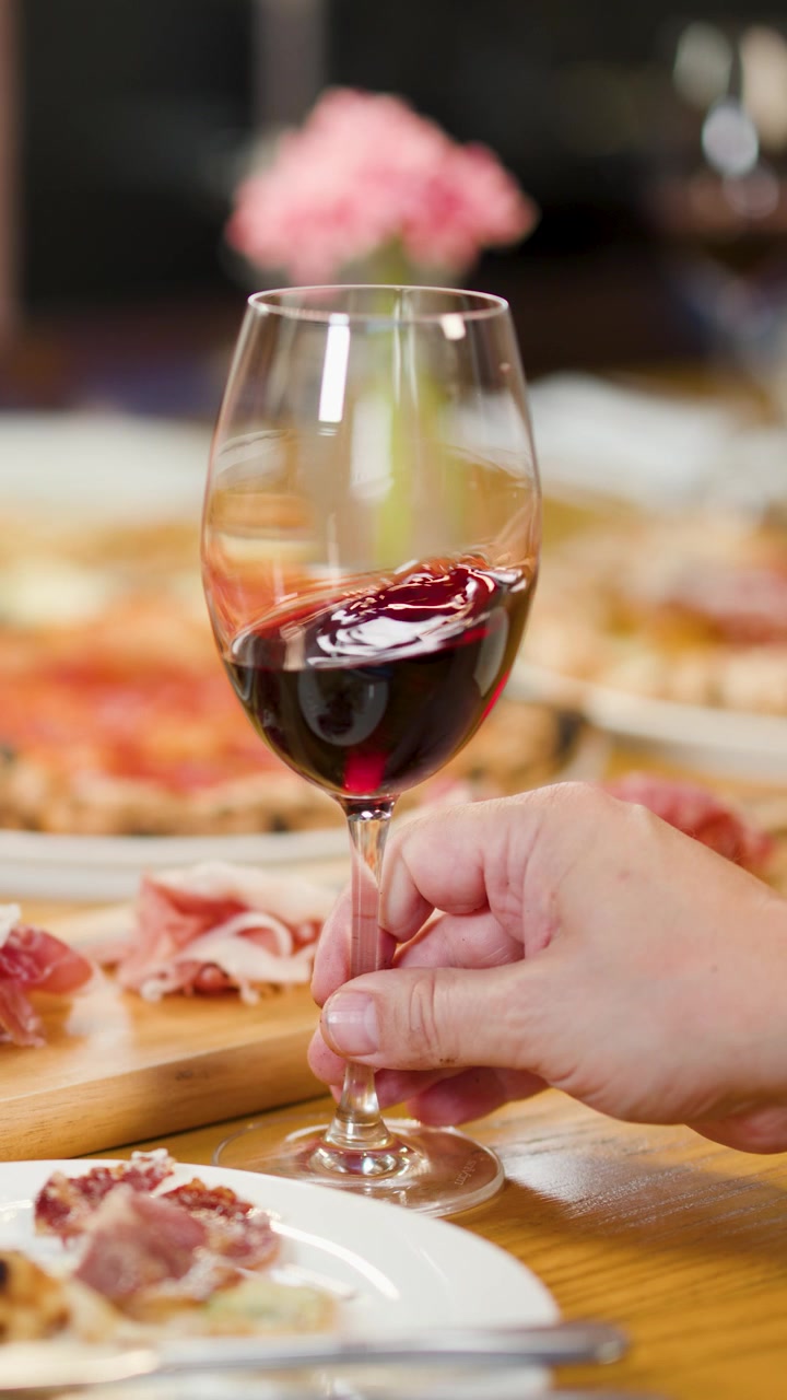 Primo piano di una mano che fa roteare vino rosso su un tavolo di legno con pizza e antipasti di prosciutto.