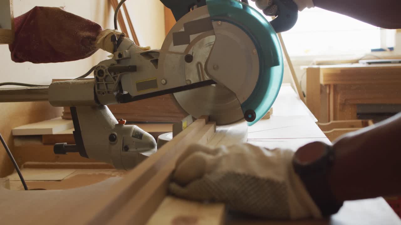 Close up of african american male carpenter hand's cutting wooden plank with electric chop saw