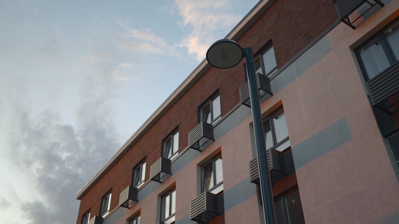 Inquadratura centrale dell'esterno di un moderno edificio residenziale con file di finestre, piccoli balconi e un lampione centrale, contro un cielo leggermente nuvoloso.