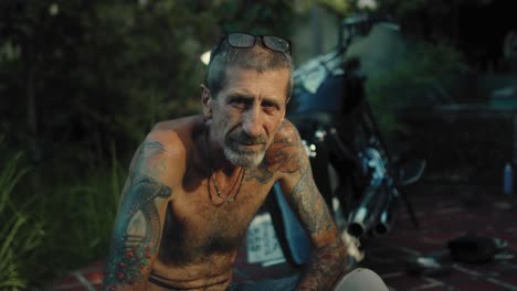 man with tattoos sitting outdoors beside a motorcycle