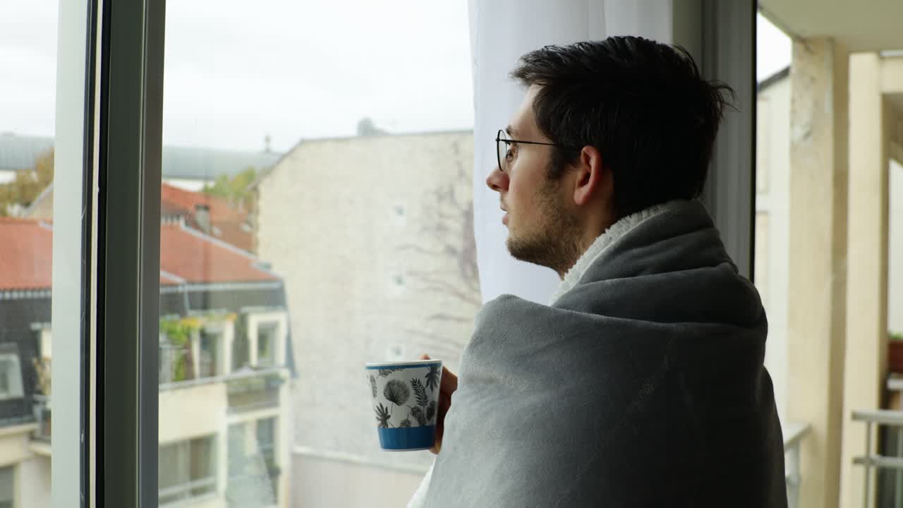 Giovane uomo che riposa a casa e guarda fuori dalla finestra in una giornata nuvolosa, con una coperta accogliente e una tazza.