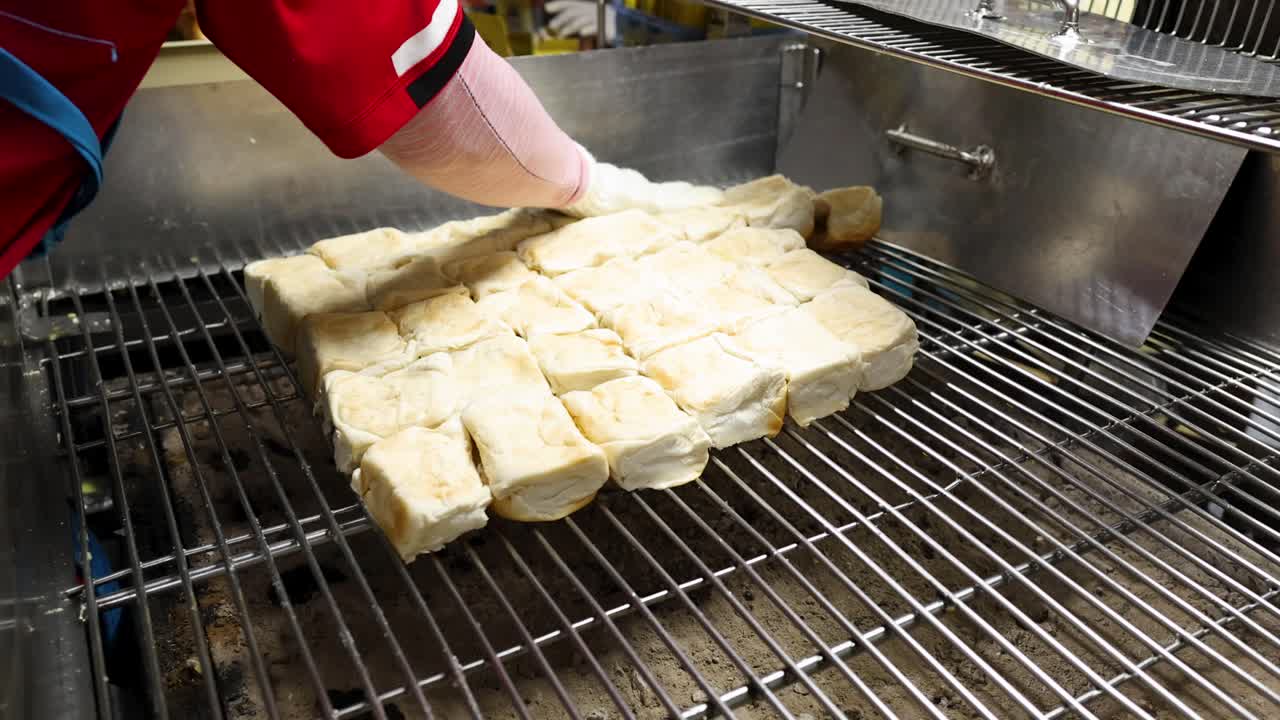 Gloved vendor toasts rows of buns on metal grill, indoor lighting, close-up, steady camera
